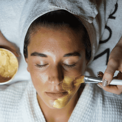 Person lying down with a brush applying a gold mask on her face